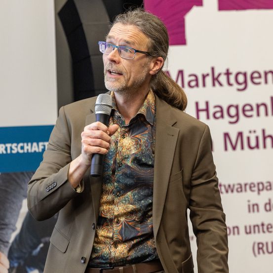 Ein Mann mit Brille und Pferdeschwanz spricht in ein Mikrofon. Er trägt ein gemustertes Hemd und einen braunen Blazer. Hinter ihm ist ein Schild mit lila und weißer Schrift.