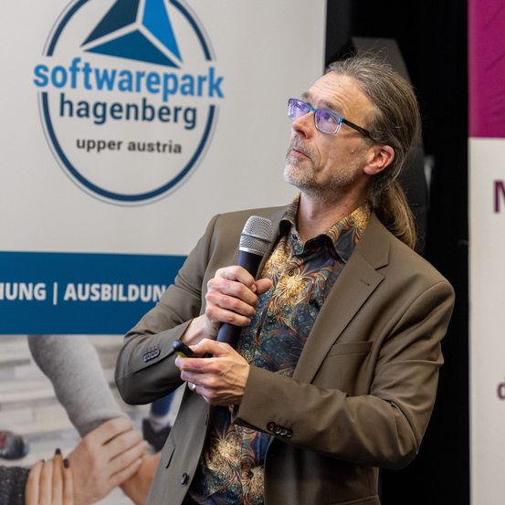 Ein Mann mit Brille und einem braunen Anzug hält ein Mikrofon in einem Konferenzraum. Hinter ihm steht ein Schild von Softwarepark Hagenberg.