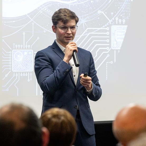 Ein Mann in einem blauen Anzug spricht in ein Mikrofon vor einer Projektionsleinwand mit Schaltkreismuster.