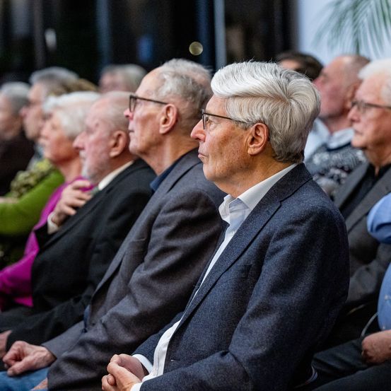 Eine Gruppe älterer Personen sitzt in einem Auditorium und sieht aufmerksam einer Präsentation zu. Der Mann im Vordergrund ist formell gekleidet, trägt eine Brille und ein weißes Hemd.