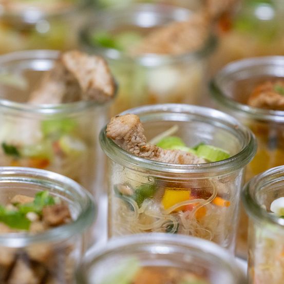 Nahaufnahme von mehreren Glasgläsern, gefüllt mit verschiedenen Lebensmitteln, einschließlich Nudeln, Gemüse und Fleisch. Die Gläser sind durchsichtig, sodass der Inhalt sichtbar ist.