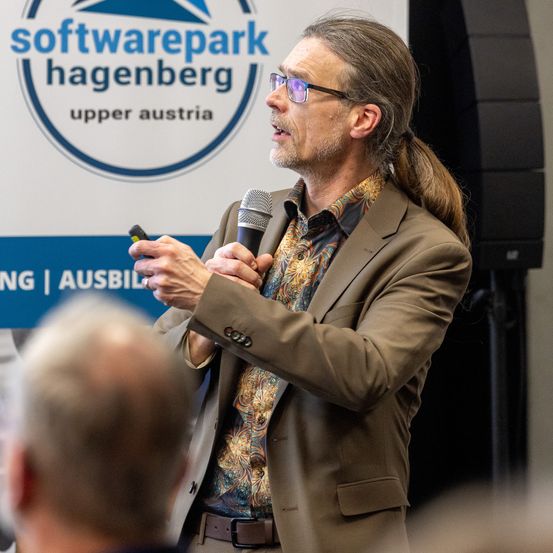 Ein erwachsener Mann mit langen Haaren und Brille spricht in ein Mikrofon. Er trägt einen braunen Anzug und ein gemustertes Hemd. Hinter ihm ist ein Schild mit dem Text Softwarepark Hagenberg.