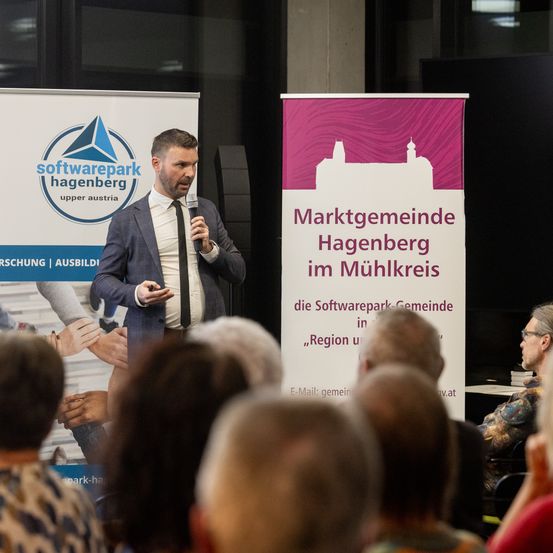 Ein Mann spricht in ein Mikrofon auf einer Konferenz, mit einem Softwarepark Hagenberg Banner im Hintergrund.
