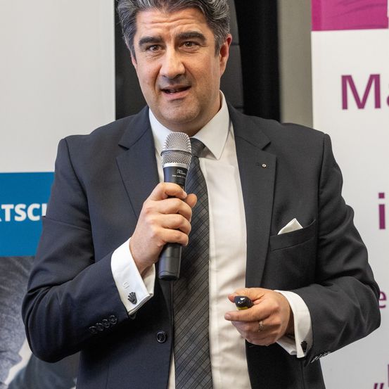 Ein Mann in einem schwarzen Anzug spricht in ein Mikrofon, vor einem Hintergrund mit einem rosa und weißen Design stehend.