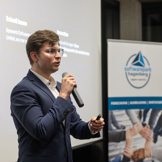 Ein Mann in Anzug und Brille hält eine Präsentation vor einer Projektionsleinwand. Er hält ein Mikrofon und einen Stift in der Hand und steht neben einem Banner.