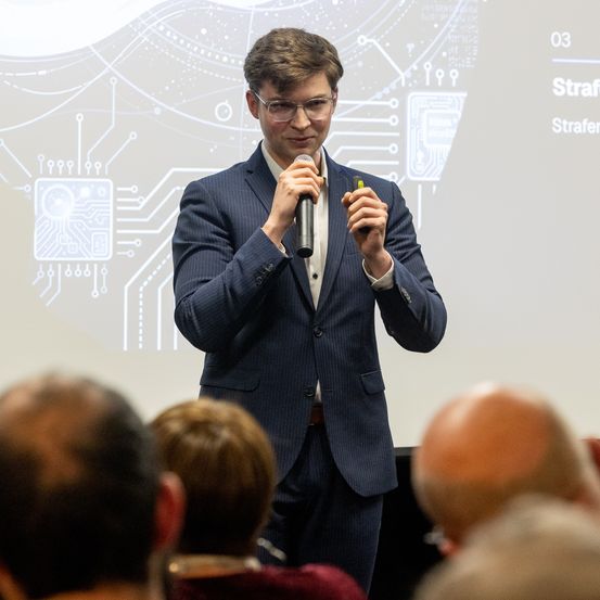 Ein Mann in einem blauen Anzug steht vor einer Projektionsleinwand und spricht in ein Mikrofon. Dahinter wird ein abstraktes Bild mit Schaltkreisen angezeigt. In der anderen Hand hält er einen grünen Marker.