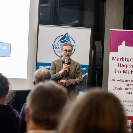 Ein Mann spricht in ein Mikrofon vor einem Publikum. Ein großer Bildschirm hinter ihm zeigt ein blaues Feld mit dem Wort 'Projekt'. Dahinter befinden sich Plakate für 'software hage' und 'Marktge Hagen im Muh'.
