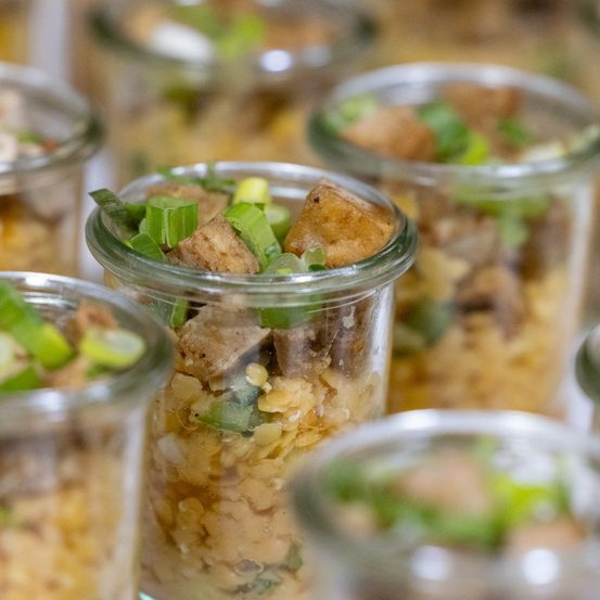 Mehrere Glasgläser mit einem Gericht gefüllt, oben mit grünen Zwiebeln und einem Stück gebratenem Tofu, auf einem Tisch präsentiert.
