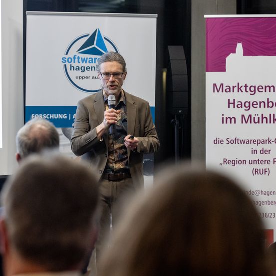 Ein Mann steht hinter einem Podium und spricht zu einem Publikum. Dahinter befindet sich ein Banner mit einem Logo für Software Hagen.