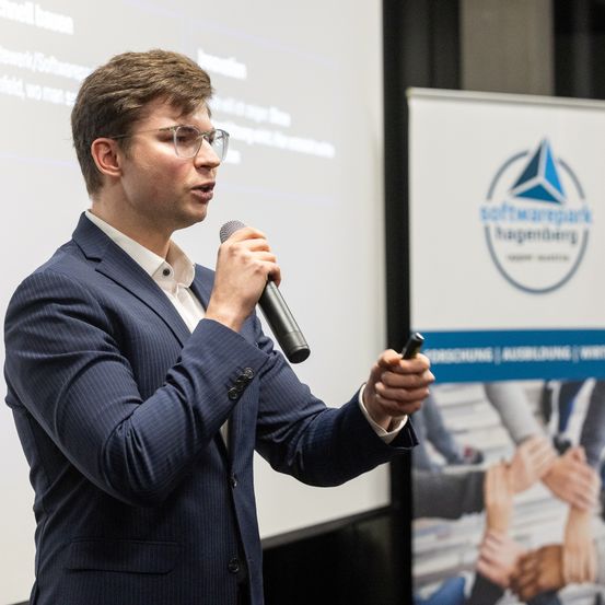 Ein Mann in einem Anzug spricht in ein Mikrofon bei einer Konferenz. Ein Banner hinter ihm hat ein Logo und Text.