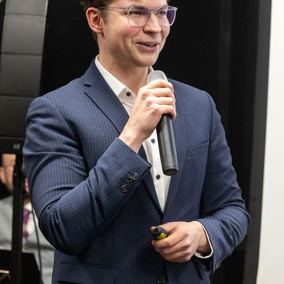 Ein Mann in einem blauen Nadelstreifenanzug lächelt und hält ein Mikrofon in der Hand. Er scheint eine Rede oder Präsentation zu halten.