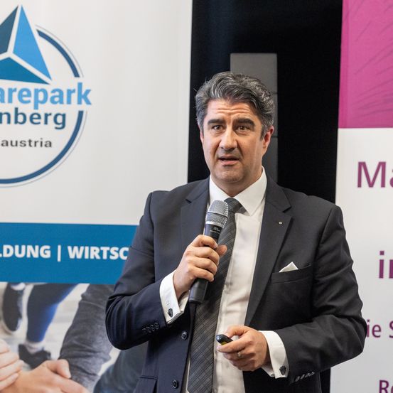 Ein Mann steht an einem Podium, hält ein Mikrofon in der Hand, trägt einen Anzug und spricht. Hinter ihm ist ein Banner mit den Worten 'Sharepark Amberg Austria.'
