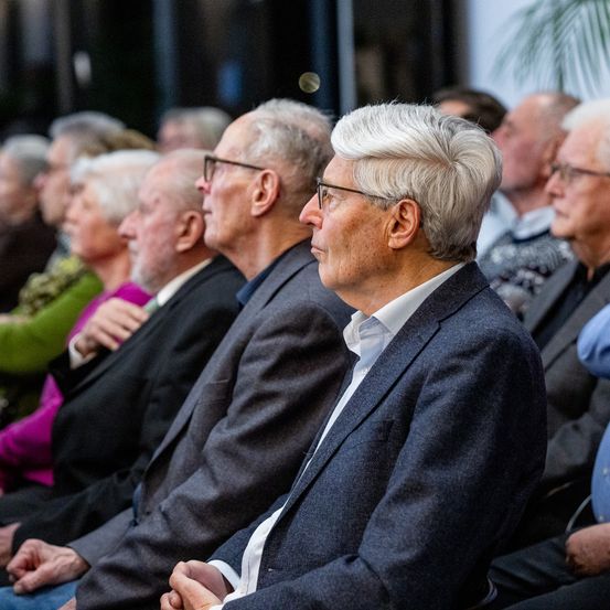 Eine Gruppe älterer Personen in formeller Kleidung sitzt in einem Auditorium und verfolgt aufmerksam eine Veranstaltung oder Präsentation.