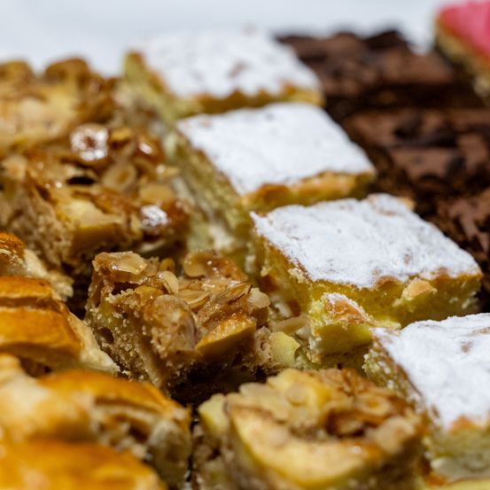 Nahaufnahme verschiedener Desserts. Verschiedene Arten von Gebäck, darunter eines mit Puderzucker, Mandelstücken und Schokolade bedeckt. Andere Desserts sind im Hintergrund sichtbar.