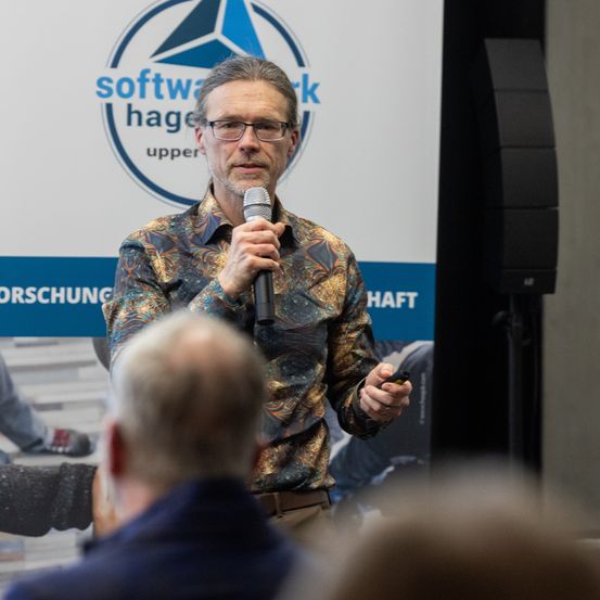 Ein Mann spricht in ein Mikrofon vor einem Publikum. Dahinter befindet sich ein Schild mit dem Text 'software hagen upper' und einem Logo.