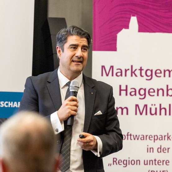 Ein Mann in einem Anzug spricht in ein Mikrofon bei einer Veranstaltung. Ein Banner mit den Worten Marktgemeinden Hagenbach im Mühlkreis in der Region darunter steht hinter ihm.