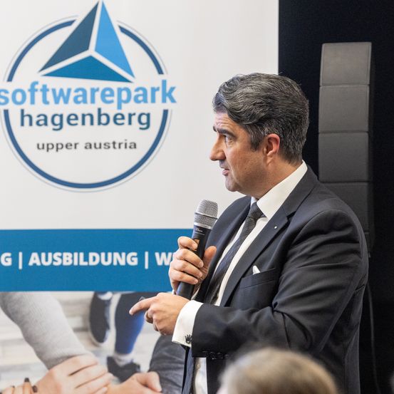 Ein Mann in einem Anzug spricht in ein Mikrofon und steht vor einem Banner mit dem Logo des Softwareparks Hagenberg in Oberösterreich.