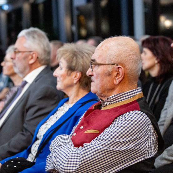 Mehrere ältere Erwachsene sitzen in einer Reihe in einem Auditorium. Ein Mann trägt eine Brille und ein kariertes Hemd.