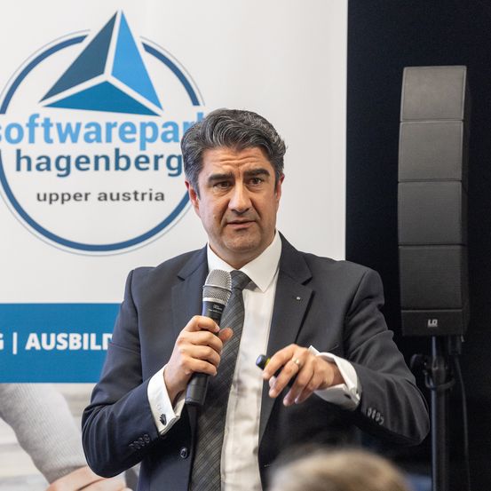 Ein Mann hält ein Mikrofon und spricht vor einem Banner für Softwarepark Hagenberg. Er trägt einen Anzug und eine Krawatte.