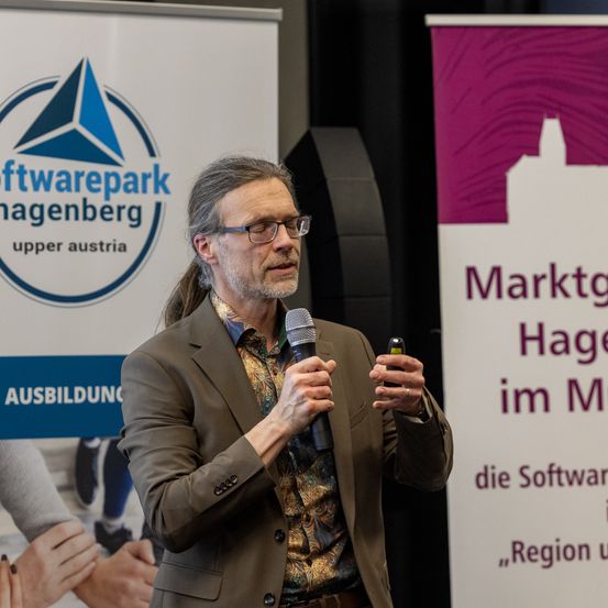 Ein Mann spricht in ein Mikrofon auf einer Konferenz vor einem Softwarepark Hagenberg Banner.