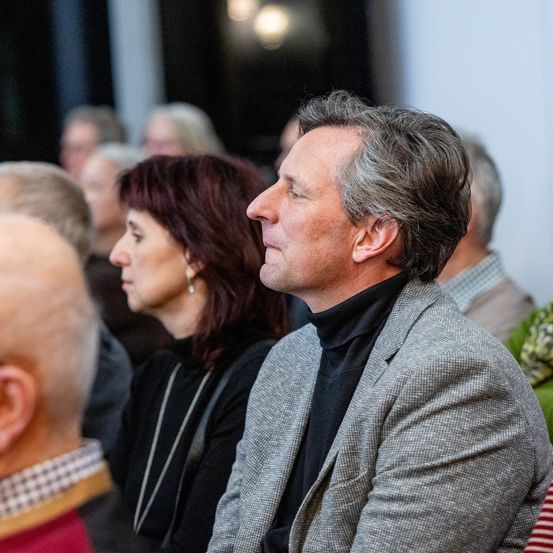 Mehrere Menschen sitzen in einem Raum, wahrscheinlich bei einer Präsentation. Ein Mann in einem grauen Anzug schaut etwas vor sich an.