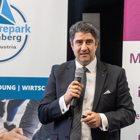 Ein Mann in Anzug und Krawatte spricht in ein Mikrofon. Hinter ihm ist eine Kulisse mit Logos und Text, von denen eines 'repark berg' lautet.