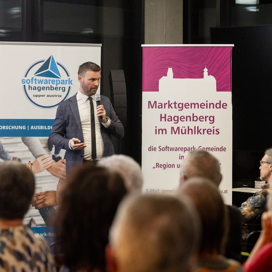 Ein Mann im Anzug spricht in ein Mikrofon vor einer Menschenmenge. Ein Plakat hinter ihm sagt 'Softwarepark Hagenberg Upper Austria'.