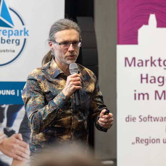 Ein Mann steht vor einem Mikrofon und spricht zu einem Publikum, mit einem Banner für 'Marktgemeinde Haging im Markt' hinter ihm.