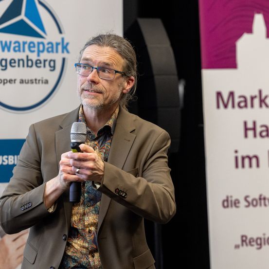 Ein Mann in einem braunen Anzug, der ein Mikrofon in der Hand hält, steht vor einem Banner mit dem Text 'Marktplatz im Haus der Software'. Er scheint bei einer Veranstaltung zu sprechen.