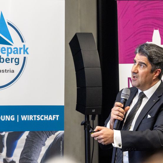 Ein Mann spricht in ein Mikrofon und hält einen Ring und ein Stück Papier in den Händen. Ein Banner mit dem Text 'wepark berg' ist auf der linken Seite. Ein Lautsprecher steht vor ihm.