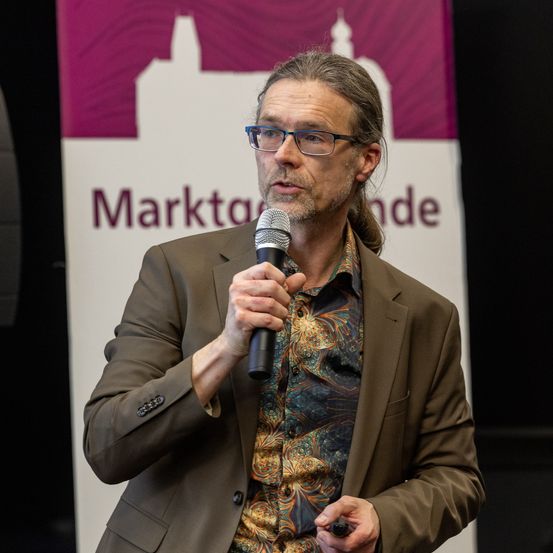 Ein Mann mit langen Haaren und Bart trägt eine Brille und spricht in ein Mikrofon vor einem Banner mit den Worten 'Marktplatz' und 'nde'.