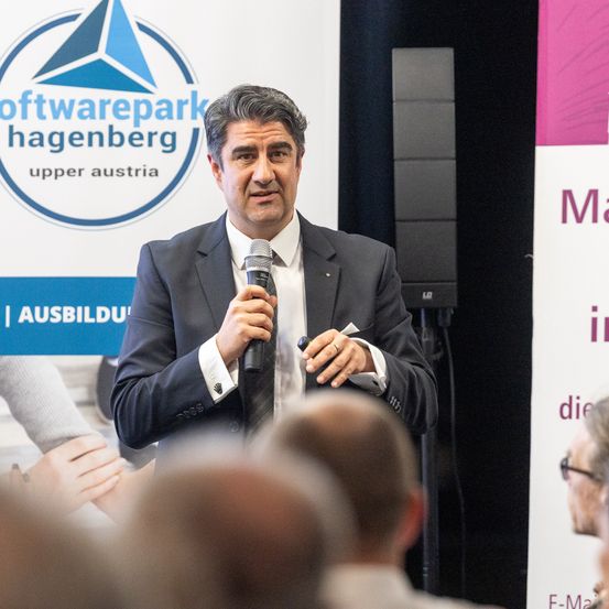 Ein Mann in einem Anzug spricht in ein Mikrofon vor einem Publikum. Hinter ihm ist ein Banner mit der Aufschrift 'Softwarepark Hagenberg Upper Austria.'