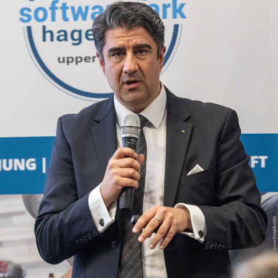 Ein Mann in einem dunklen Anzug und Krawatte steht und spricht in ein Mikrofon bei einer Veranstaltung. Hinter ihm ist ein Banner mit dem Text 'Software Hage' und 'upper'.