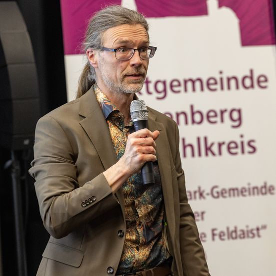 Ein Mann mit Brille und Bart, der ein Mikrofon hält, scheint eine Rede bei einer Veranstaltung zu halten. Hinter ihm ist ein Banner mit Text auf Deutsch.