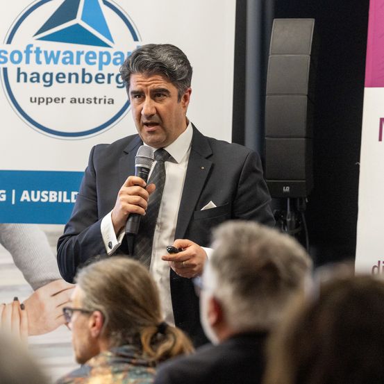 Ein Mann in einem formellen Anzug spricht ein Publikum mit einem Mikrofon an. Hinter ihm befindet sich ein Hintergrund mit dem Text 'Softwarepark Hagenberg' und 'Oberösterreich'.