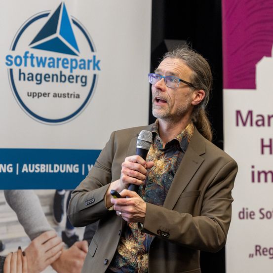 Ein Mann in einem braunen Anzug spricht in ein Mikrofon auf einer Konferenz im Softwarepark Hagenberg in Oberösterreich.