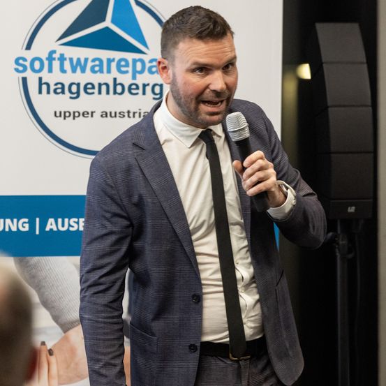 Ein Mann in dunklem Anzug und Krawatte spricht in ein Mikrofon vor einem Banner mit den Worten 'Softwarepa Hagenberg Upper Austria'.