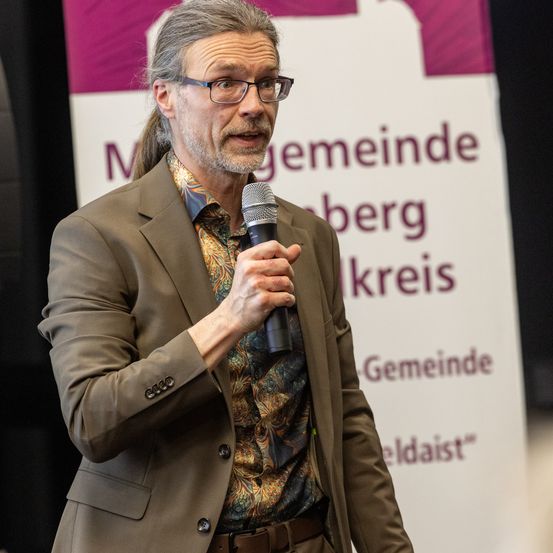 Ein Mann in einem Anzug und mit Brille spricht in ein Mikrofon vor einem Schild.