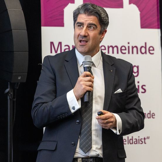 Ein Mann in Anzug und Krawatte hält ein Mikrofon und spricht bei einer Veranstaltung. Hinter ihm steht ein Banner mit der Aufschrift 'Marxemeinde'.