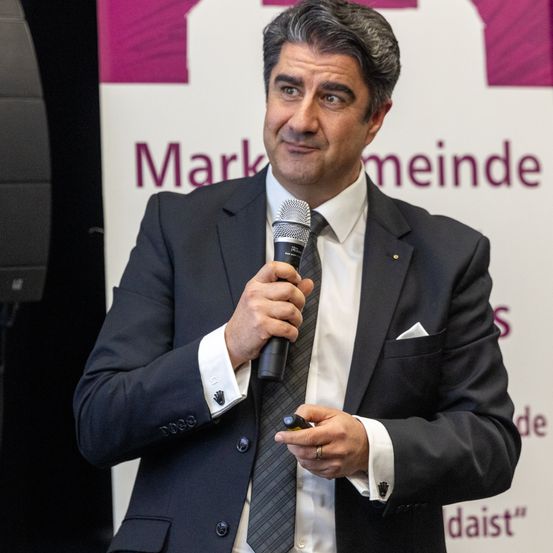 Ein Mann in einem formellen Anzug hält ein Mikrofon und scheint bei einer Veranstaltung zu sprechen. Hinter ihm befindet sich ein Banner mit dem Text 'Marktgemeinde'.