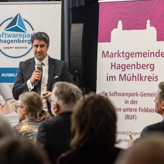 Ein Mann spricht mit einem Mikrofon vor einer Menschenmenge in einem Raum, mit einem Softwarepark Hagenberg-Banner im Hintergrund.