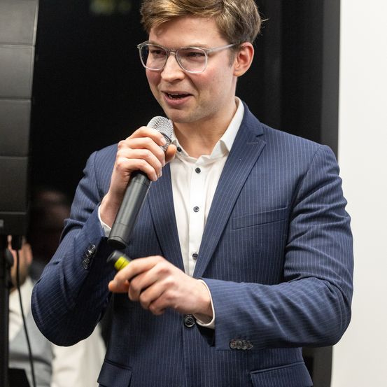 Ein Mann in einem blauen Anzug hält ein Mikrofon und spricht bei einer Veranstaltung. Er trägt eine Brille und ein weißes Hemd. Der Hintergrund ist unscharf mit einer dunklen Wand.