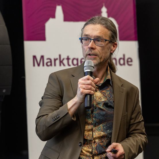 Ein Mann mit Brille und einem braunen Anzug spricht in ein Mikrofon, möglicherweise bei einer Veranstaltung. Hinter ihm befindet sich ein Banner mit dem Text 'Marktga de'.