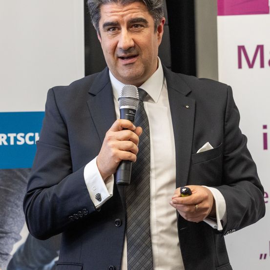 Ein Mann in formeller Kleidung hält ein Mikrofon und spricht bei einer Veranstaltung. Er trägt eine Krawatte und einen Ring am Finger.