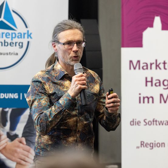 Ein Mann in einem gemusterten Hemd spricht in ein Mikrofon und hält einen kleinen Gegenstand in der anderen Hand vor einem Schild mit Logo und Text.