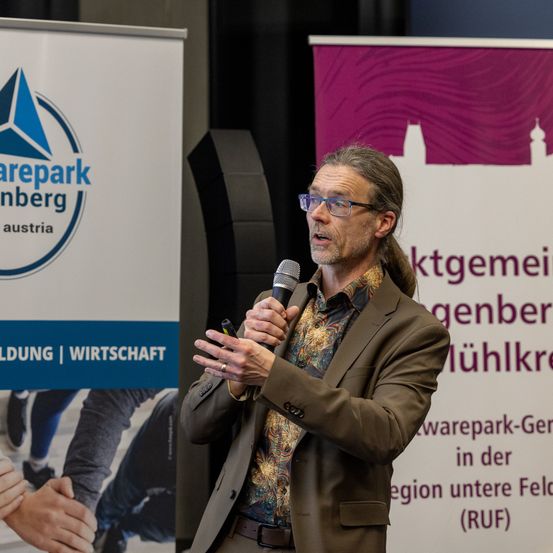 Ein Mann spricht in ein Mikrofon bei einer Veranstaltung. Hinter ihm ist ein Banner mit einem Logo und Text. Links ist eine Figur zu sehen, die Hände schüttelt.