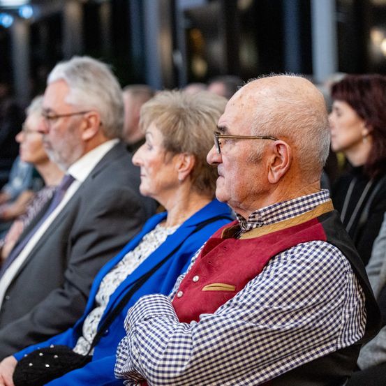 Eine Gruppe älterer Personen sitzt in einem Theater und sieht sich eine Aufführung an. Ein Mann in einem karierten Hemd schaut nach vorne, während ein anderer Mann in einem Anzug ihn ansieht.