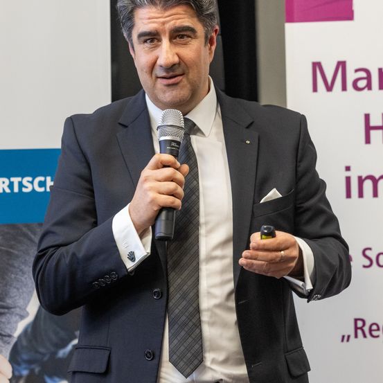 Ein Mann in einem dunklen Anzug, der ein Mikrofon hält und lächelnd hineinspricht, möglicherweise bei einer Veranstaltung.