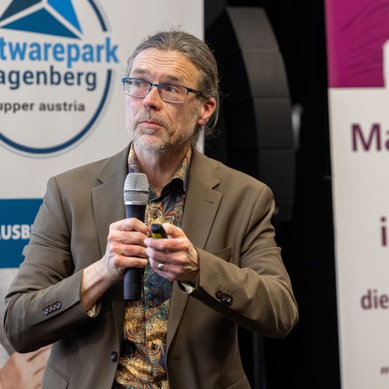 Ein Mann in einem braunen Anzug hält ein Mikrofon und spricht vor einem Schild des 'Softwarepark'.