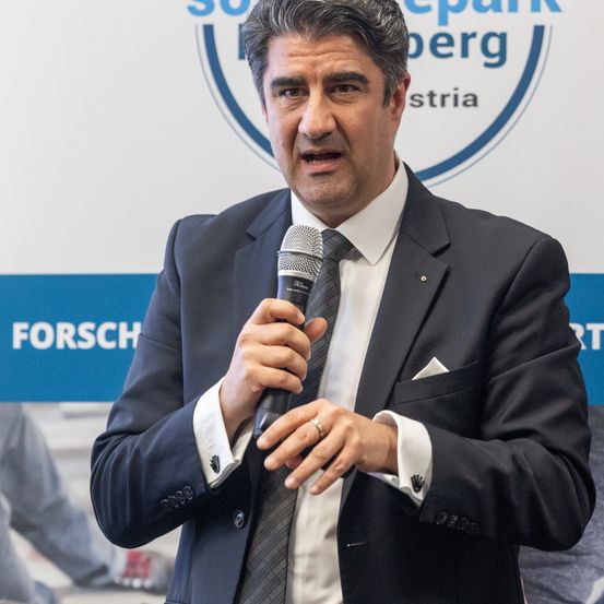 Ein Mann in einem dunklen Anzug spricht in ein Mikrofon auf einer Konferenz. Der Hintergrund zeigt ein Logo mit Text.